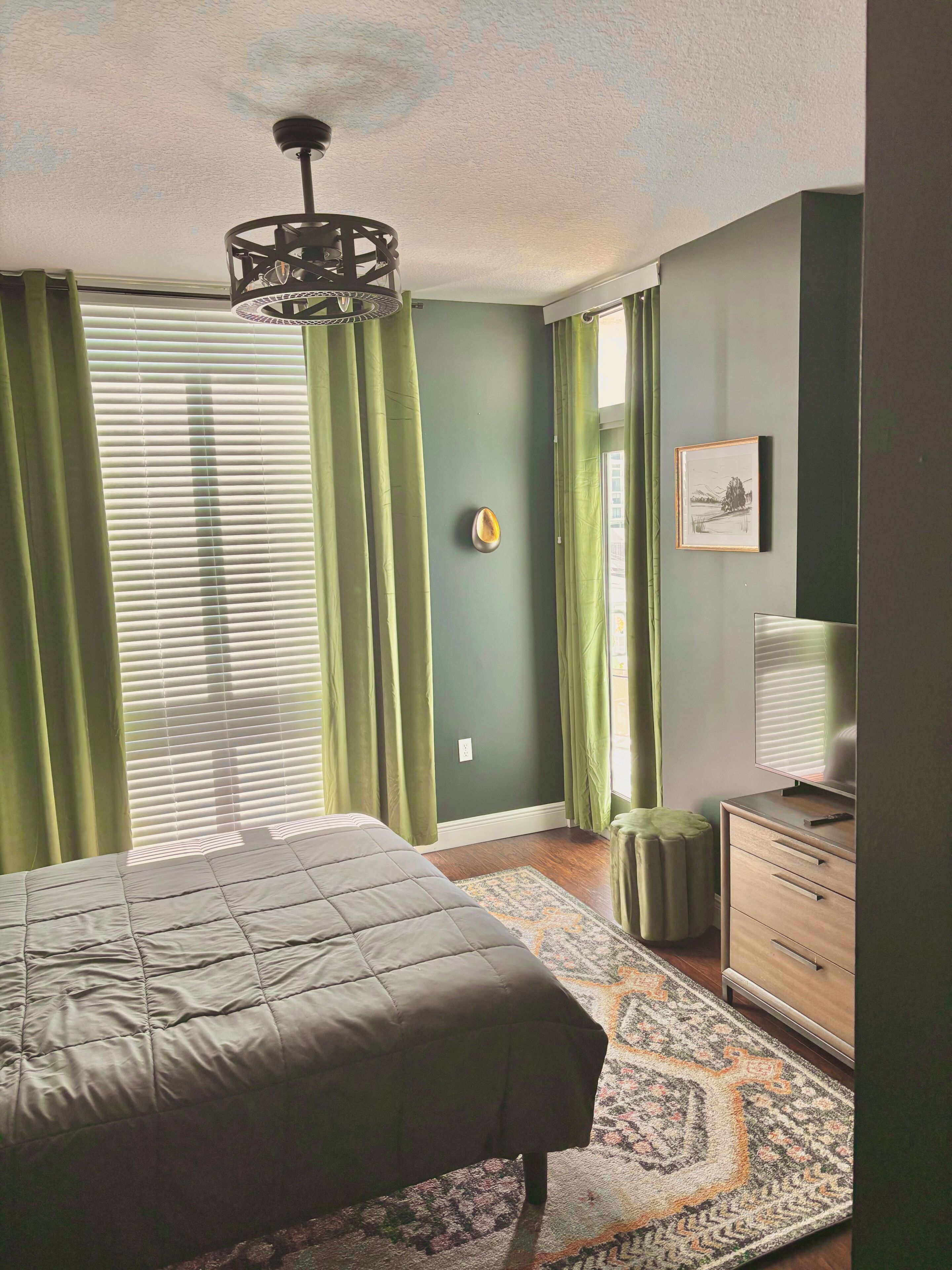 A bedroom with a bed, a dresser, a lamp, a wall-mounted heater, and a window with curtains.
