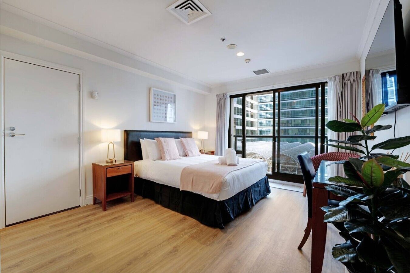 A hotel room with a bed, bedside table, lamp, window with curtains, and a plant.