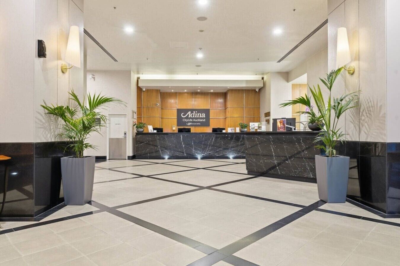 Een hotel receptie met een marmeren balie, plantenpotten en een bord met de tekst 'Adina CityLife Auckland'.