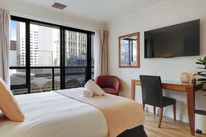 A hotel room with a bed, a desk, a chair, a TV, and a window with a view of buildings. - Queen Street Gem - Freshly Renovated Studio (Auckland CBD)