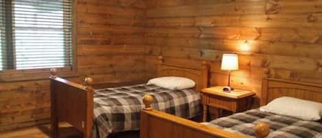 A cabin-style room with two wooden beds, a nightstand with a lamp, and a window with blinds.