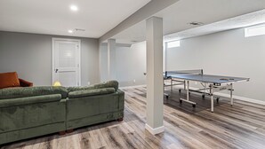 A modern living room with a green sofa, a ping pong table, and a door with louvered panels. - Amber & Oak: Theater, Game Room & Fire Pit (Omaha)