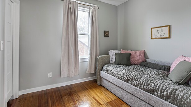A room with a single bed, a ceiling fan, a window with curtains, and a wall-mounted picture. - Amber & Oak: Theater, Game Room & Fire Pit (Omaha)