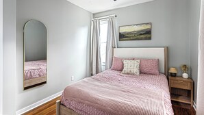 A bedroom with a bed, a nightstand, a lamp, a mirror, a ceiling fan, and a painting on the wall. - Amber & Oak: Theater, Game Room & Fire Pit (Omaha)