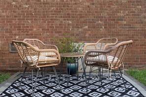 A patio with two wicker chairs and a small table, a potted plant, and a patterned rug against a brick wall. - Renovated 3BR House - Monthly Stays - Fenced Quarter-Acre Yard - Pets Welcome (Charlotte)