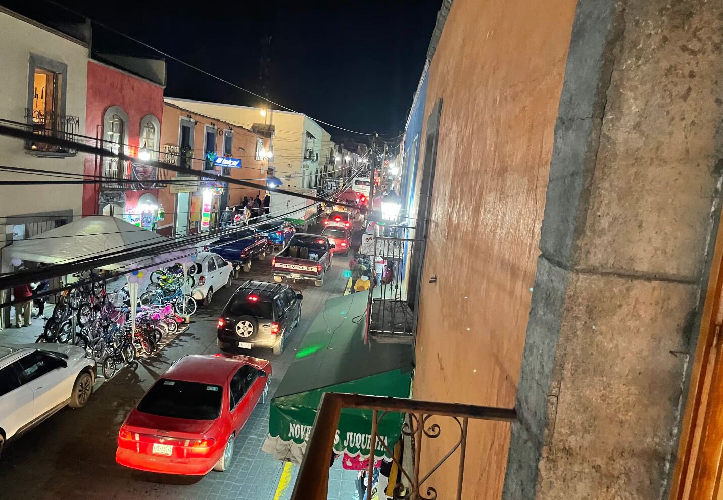 Escena de carrer de nit amb cotxes i motos aparcades, edificis amb cartelleria i un local comercial visible.