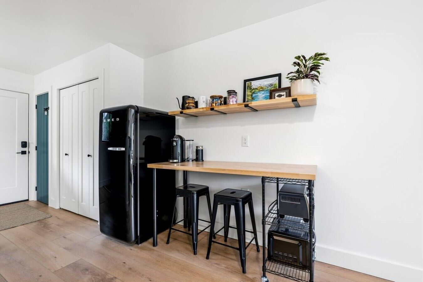 A compact kitchen with a black refrigerator, a wooden countertop, two black stools, and a small shelf with decorative items.