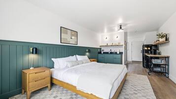 A bedroom with a bed, bedside tables, a nightstand, a wall-mounted lamp, a framed picture, and a kitchenette area with a countertop, sink, and appliances.