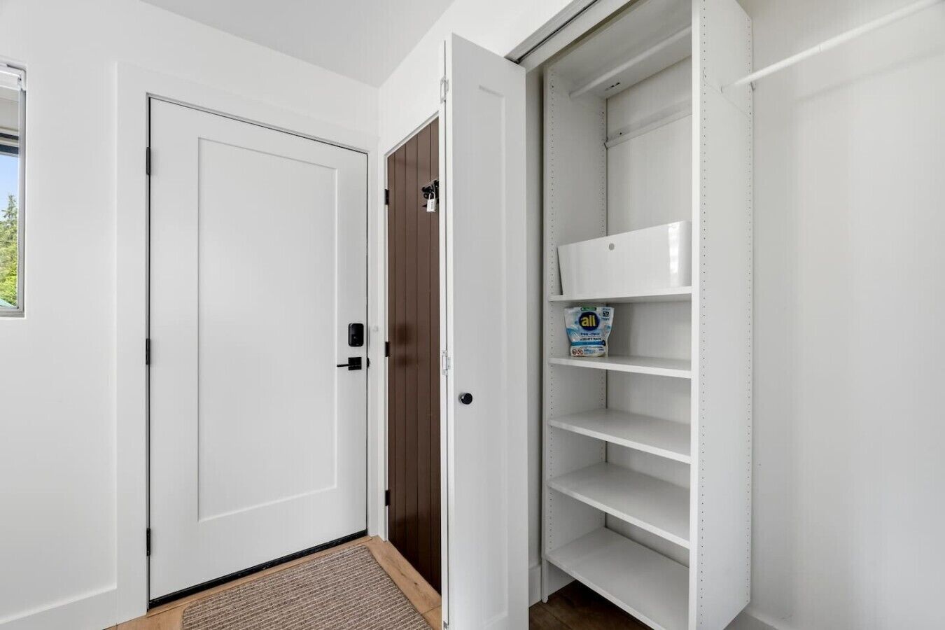 Un couloir avec une porte blanche, une porte marron et une étagère blanche contenant une boîte de lessive.