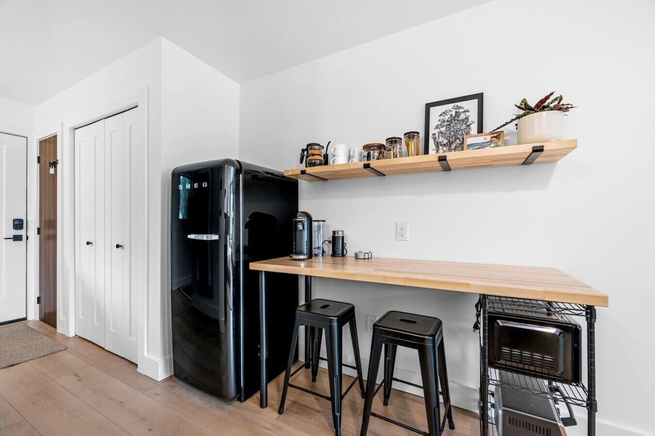 Une cuisine compacte avec un réfrigérateur noir, un plan de travail en bois, deux tabourets, une machine à café et un four à micro-ondes.