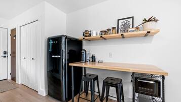Eine kleine Küche mit einem schwarzen Kühlschrank, einer Holzarbeitsplatte, zwei Barhocker, einer Kaffeemaschine und einer Mikrowelle.