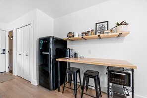 Une cuisine compacte avec un réfrigérateur noir, un plan de travail en bois, deux tabourets, une machine à café et un four à micro-ondes.