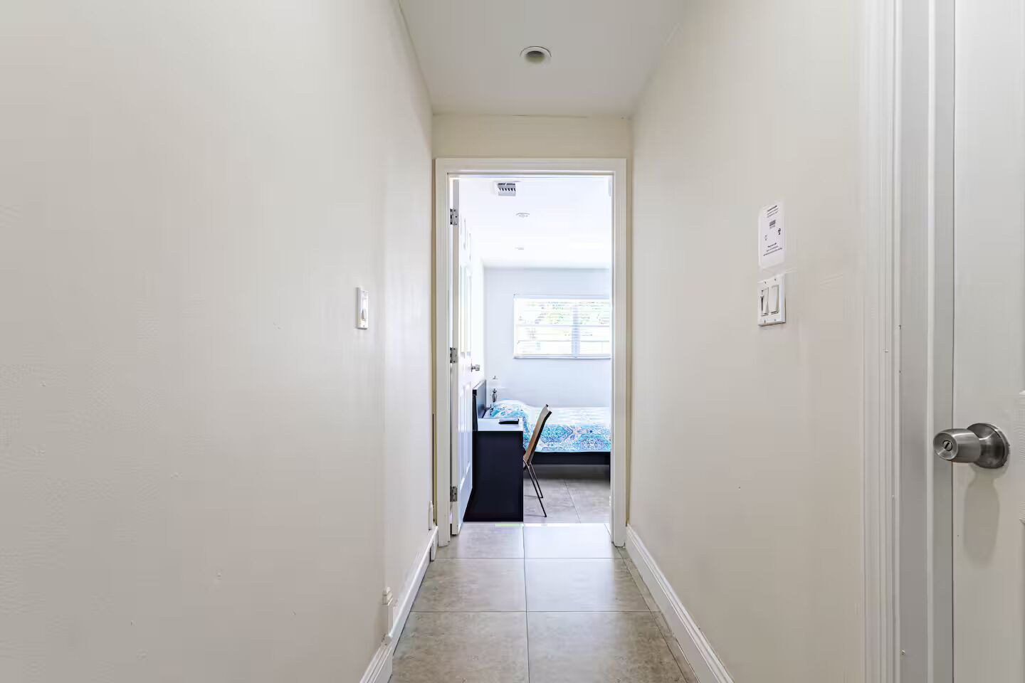 A narrow hallway with a door on the right, leading to a room with a bed and a desk.