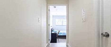 A narrow hallway with a door on the right, leading to a room with a bed and a desk.