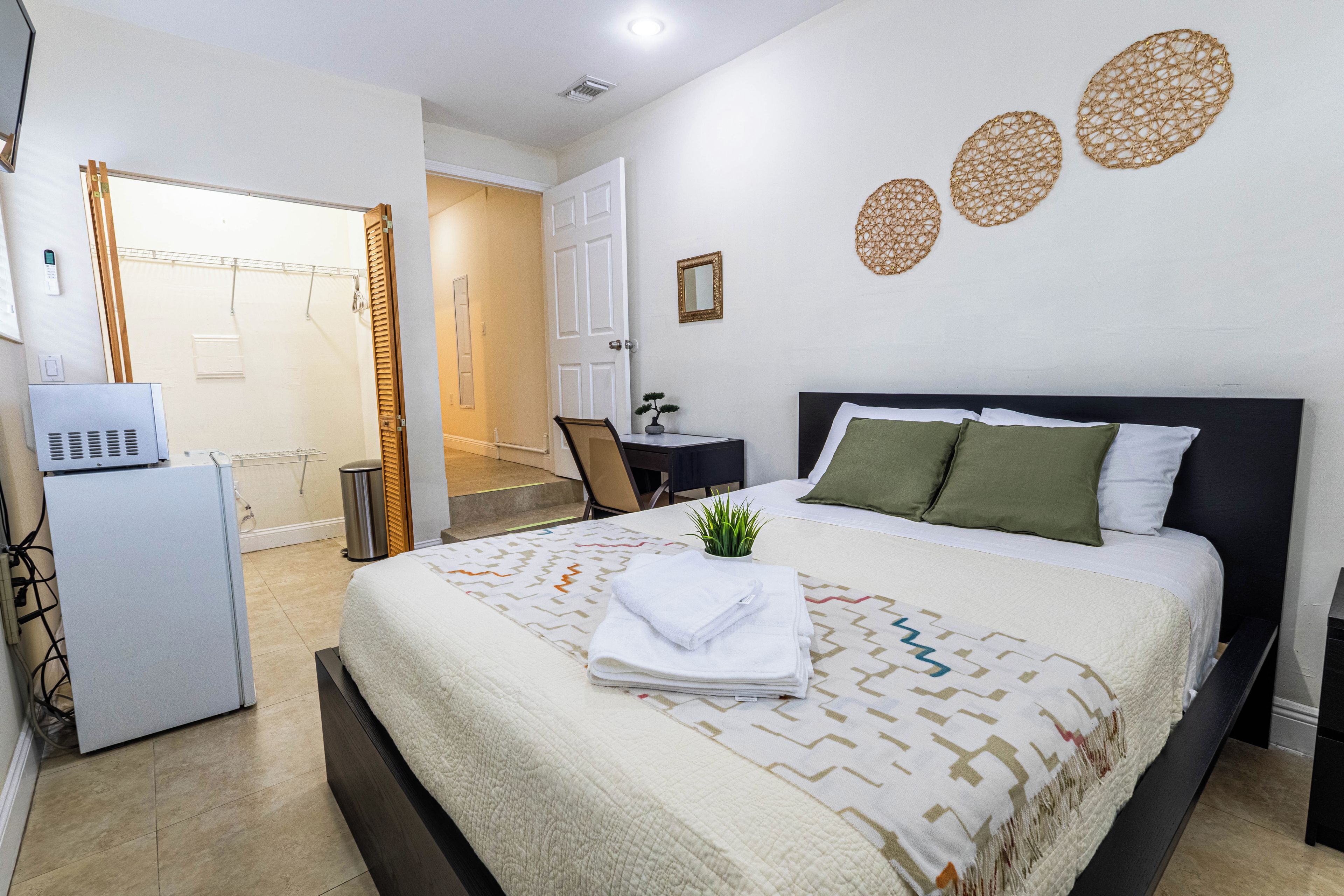 A hotel room with a bed, a desk, a chair, a small plant, and a mirror on the wall.
