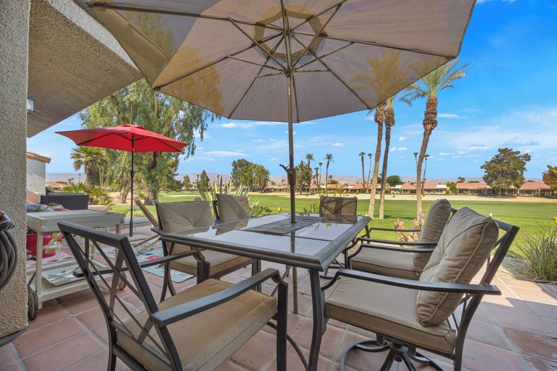 An outdoor patio with a table and chairs, a red umbrella, and a view of a golf course.