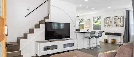 A modern living room with a flat-screen TV, a staircase, a dining area with bar stools, and artwork on the walls.