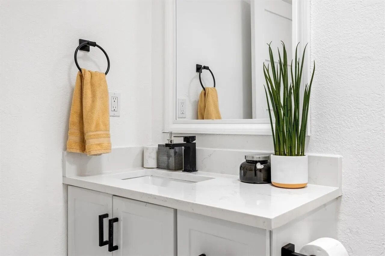 Ein Badezimmer mit einem weißen Waschbecken, einem Spiegel, einem Handtuchhalter mit einem gelben Handtuch, einer Topfpflanze und einem Toilettenpapierhalter.