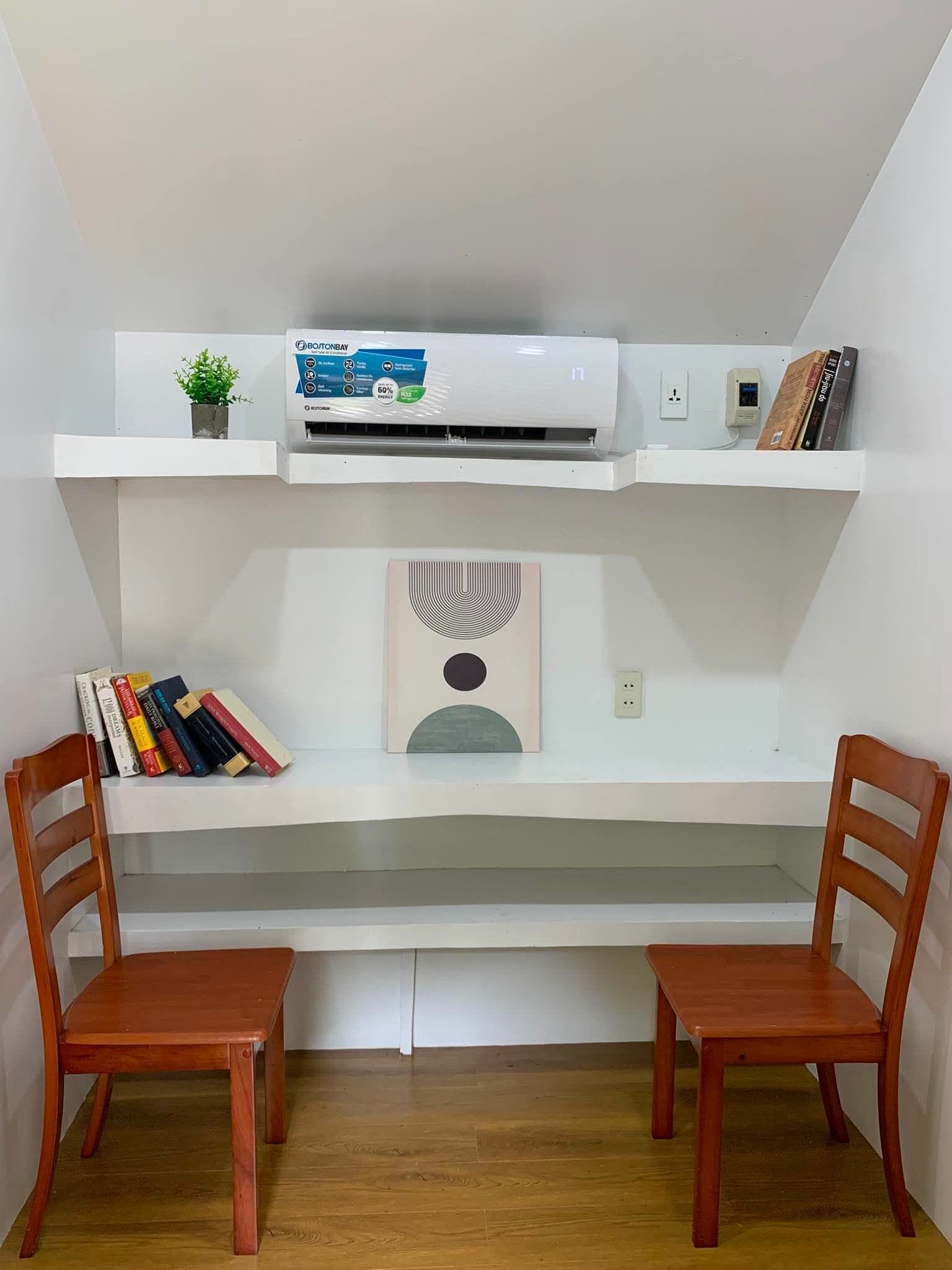 Un espace de travail comprenant un bureau, deux chaises, une bibliothèque, un climatiseur et une étagère murale agrémentée d’une plante et de livres.