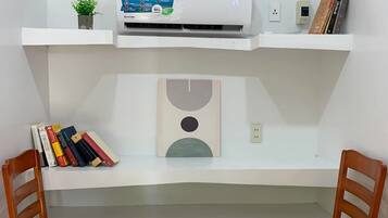 Un espace de travail comprenant un bureau, deux chaises, une bibliothèque, un climatiseur et une étagère murale agrémentée d’une plante et de livres.
