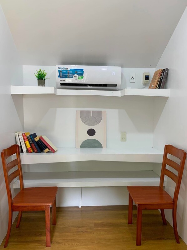 A study area with a desk, two chairs, a bookshelf, an air conditioner, and a wall-mounted shelf with a plant and books.
