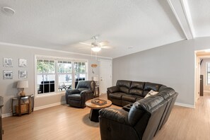 A spacious living room with a large leather sofa, a smaller armchair, and a coffee table. There are large windows providing natural light and a view of the outdoors. - Lake it Easy Private Dock with fish cleaning station (Batesburg-Leesville)