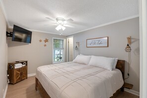 Une chambre à coucher comprenant un lit, un ventilateur de plafond, un téléviseur fixé au mur, une armoire en bois et une lampe fixée au mur.
