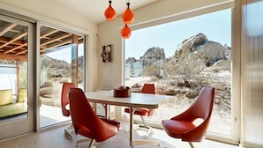 A modern interior with a dining area, red chairs, a table, and a view of rocky terrain. - Radziner Modernist Cabin By Homestead Modern (Joshua Tree)