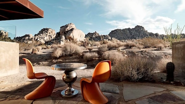 An outdoor seating area with two orange chairs and a table, set against a desert landscape with rocky formations and sparse vegetation. - Radziner Modernist Cabin By Homestead Modern (Joshua Tree)