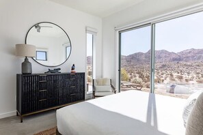 A bedroom with a large bed, a dresser, a mirror, and a view of a desert landscape through a sliding glass door. - Shelter 2 By Homestead Modern (Joshua Tree)