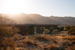 Ein modernes Haus mit Flachdach, einer kleinen Veranda mit Stühlen und Blick auf Berge im Hintergrund.
