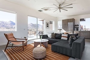 Ein modernes Wohnzimmer mit Sofa, Sessel und Couchtisch, einem Küchenbereich und großen Fenstern mit Blick auf die Berge.