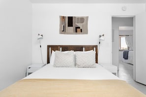 Quarto com cabeceira de madeira, cama com roupa de cama branca, duas luminárias de cabeceira e uma obra de arte abstrata na parede.