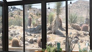 A conservatory with a view of desert cacti, wooden furniture, and a white table. - Sommer Ranch By Homestead Modern (Yucca Valley)