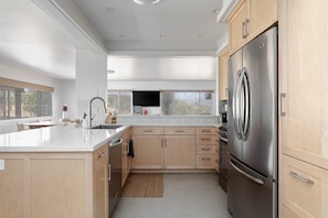 A modern kitchen with a large stainless steel refrigerator, wooden cabinets, a white countertop, and a window providing a view of the outdoors. - Sommer Ranch By Homestead Modern (Yucca Valley)