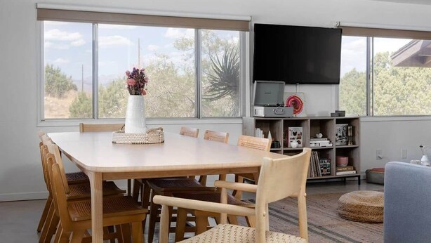 A dining area with a large table and chairs, a television mounted on the wall, and a bookshelf with various items. - Sommer Ranch By Homestead Modern (Yucca Valley)