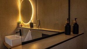 A modern bathroom with a rectangular sink, a large mirror with a circular light, and two black bottles on the countertop. - Iuman Patagonia (El Calafate)