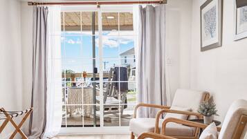 A room with a sliding glass door leading to an outdoor deck, a cushioned chair, a small table with a vase, and a framed picture on the wall.