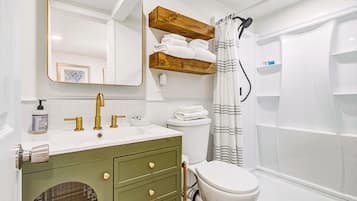 Ein Badezimmer mit einem weißen Waschbecken, goldenen Wasserhähnen, einer grünen Waschtischablage, einer Toilette, einer Dusche mit gestreifter Duschvorhang und einem Holzregal mit gefalteten Handtüchern.