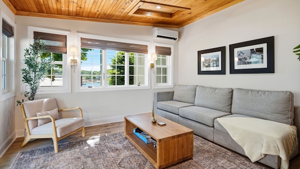 A living room with a wooden ceiling, a grey sofa, a wooden coffee table, a patterned rug, a plant, and two framed pictures on the wall.