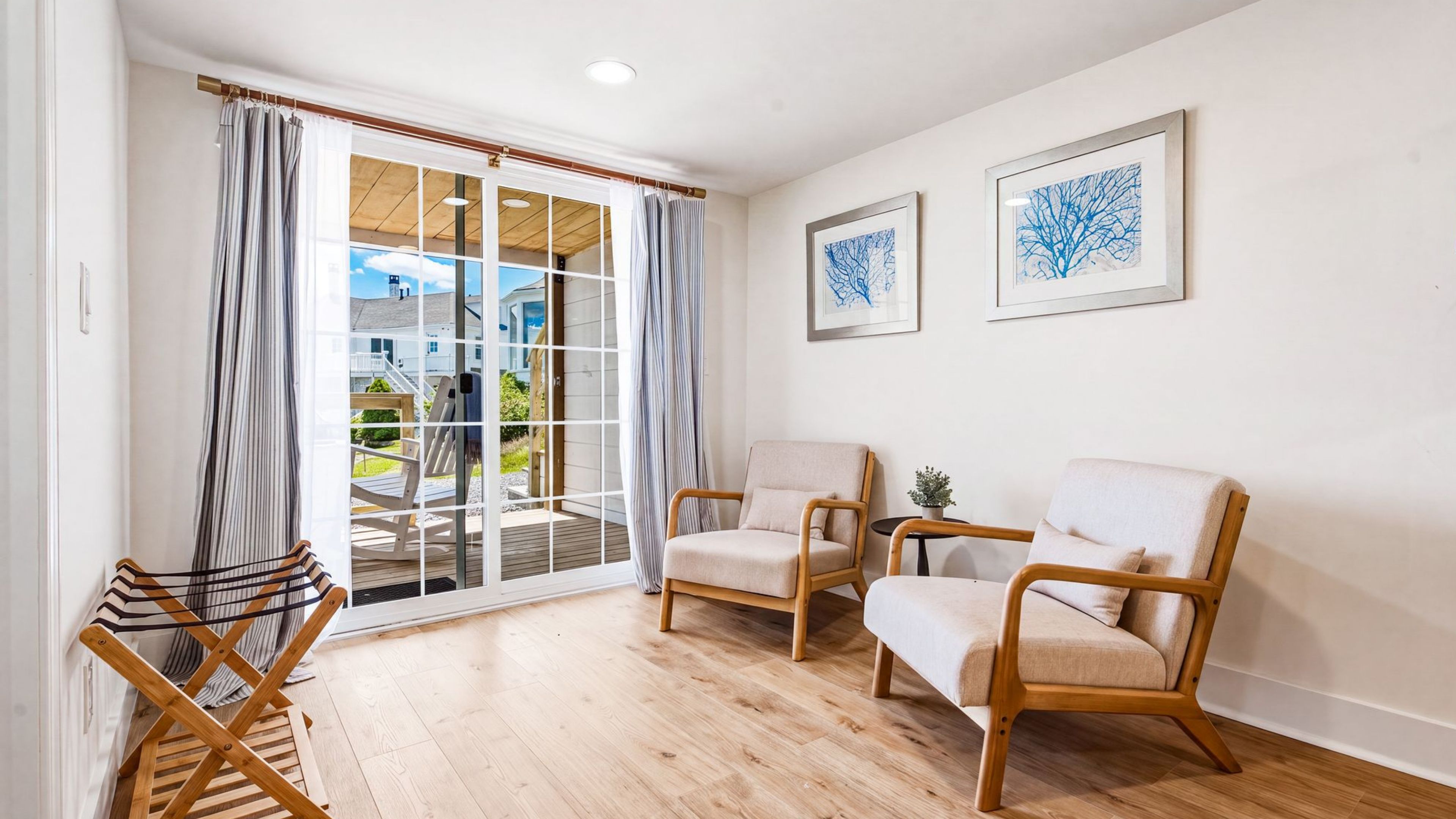 Ein Zimmer mit Holzboden, zwei beigefarbene Sessel, ein kleiner Tisch mit einer Pflanze und Blick durch eine Glastür auf einen Balkon.
