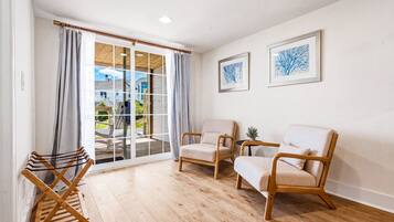 Ein Zimmer mit Holzboden, zwei beigefarbene Sessel, ein kleiner Tisch mit einer Pflanze und Blick durch eine Glastür auf einen Balkon.