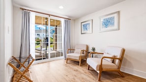 A room with a wooden floor, two beige armchairs, a small table with a plant, and a view through a glass door to a balcony.