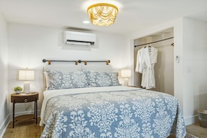 A bedroom with a bed, bedside lamps, an air conditioning unit, a robe hanging, and a shower area.