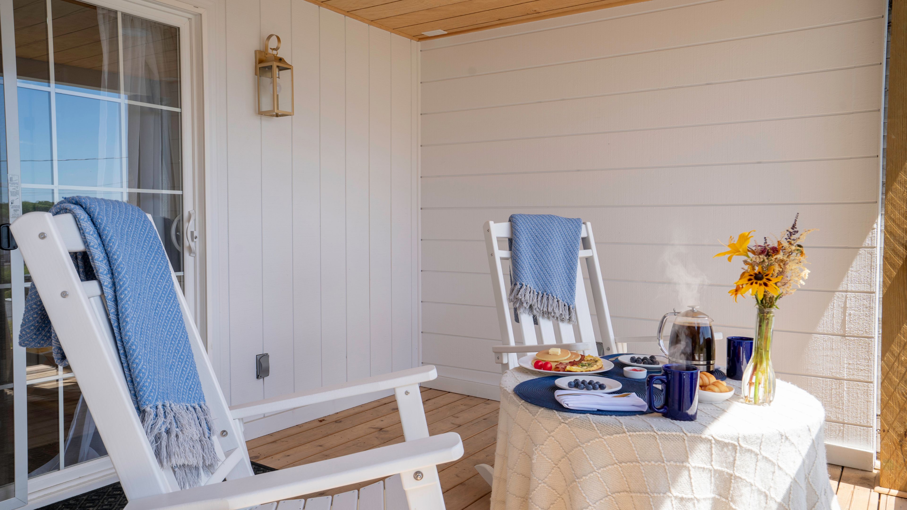 A covered outdoor seating area with two white chairs, a table with breakfast items, and a vase with flowers.
