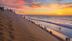 Een strand met mensen die langs de waterlijn wandelen en een ondergaande zon op de achtergrond.