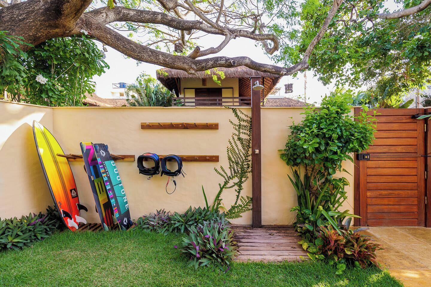 Un jardin entouré d’une clôture en bois, une construction à toit de chaume et deux planches de surf appuyées contre le mur.
