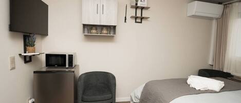 A hotel room with a bed, a desk with a microwave, a chair, a TV mounted on the wall, and a small shelf with decorative items.