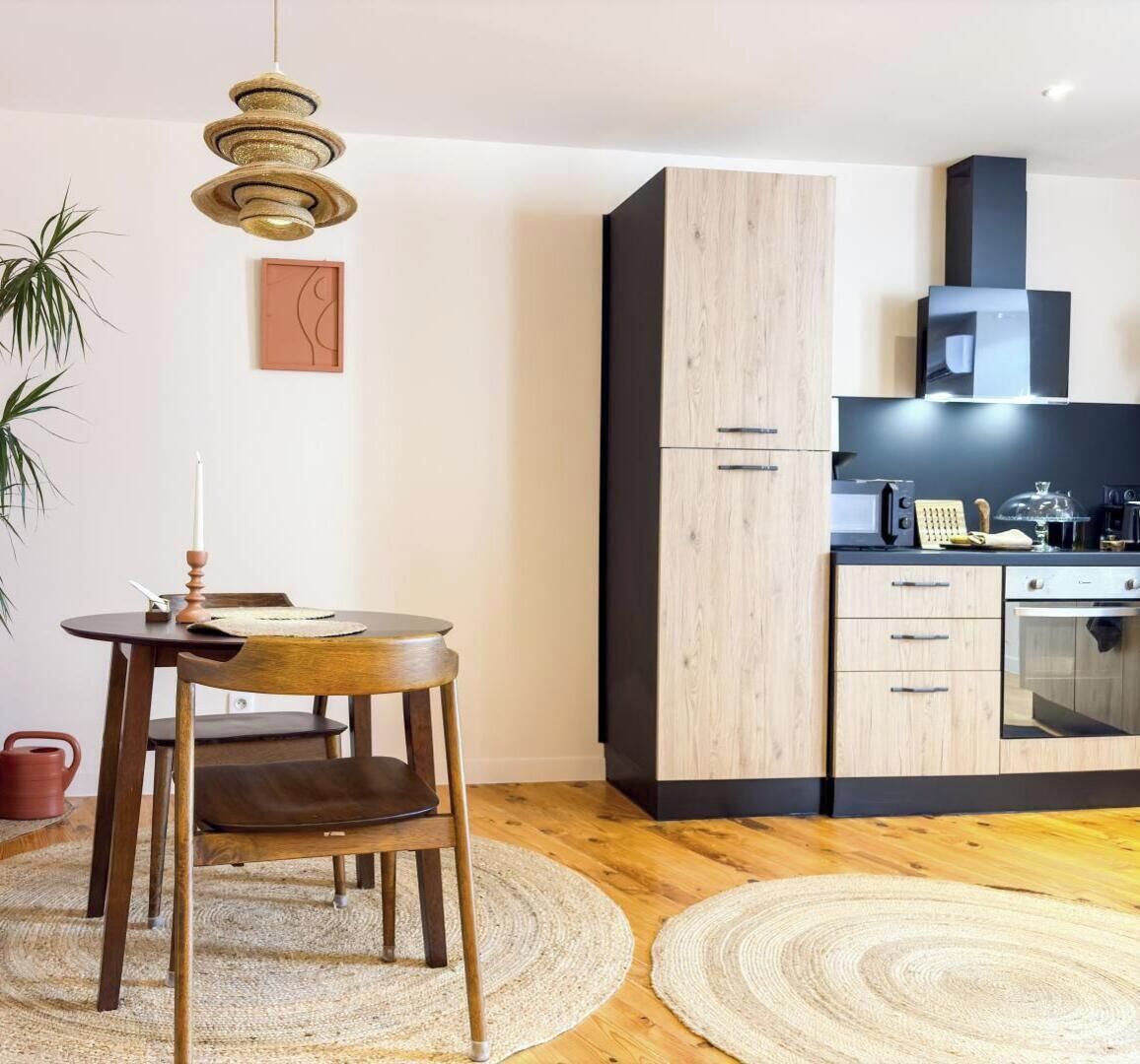 Een moderne keuken met houten kastjes, een eethoek met een ronde tafel en stoelen, en een hangende lamp.