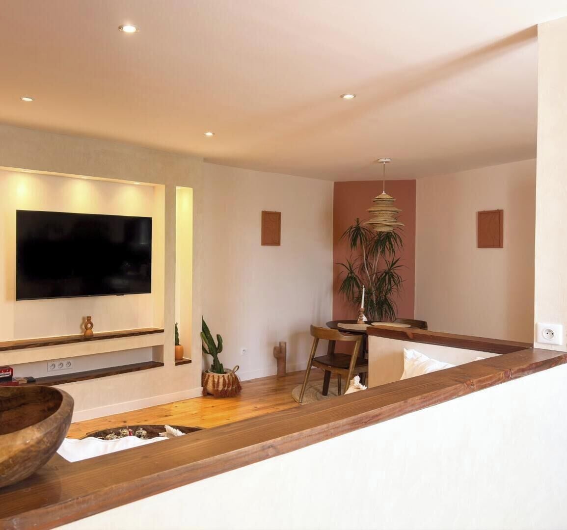 Una sala moderna con televisor de pantalla plana montado en la pared, un comedor con mesa y sillas de madera, y una planta grande en maceta.
